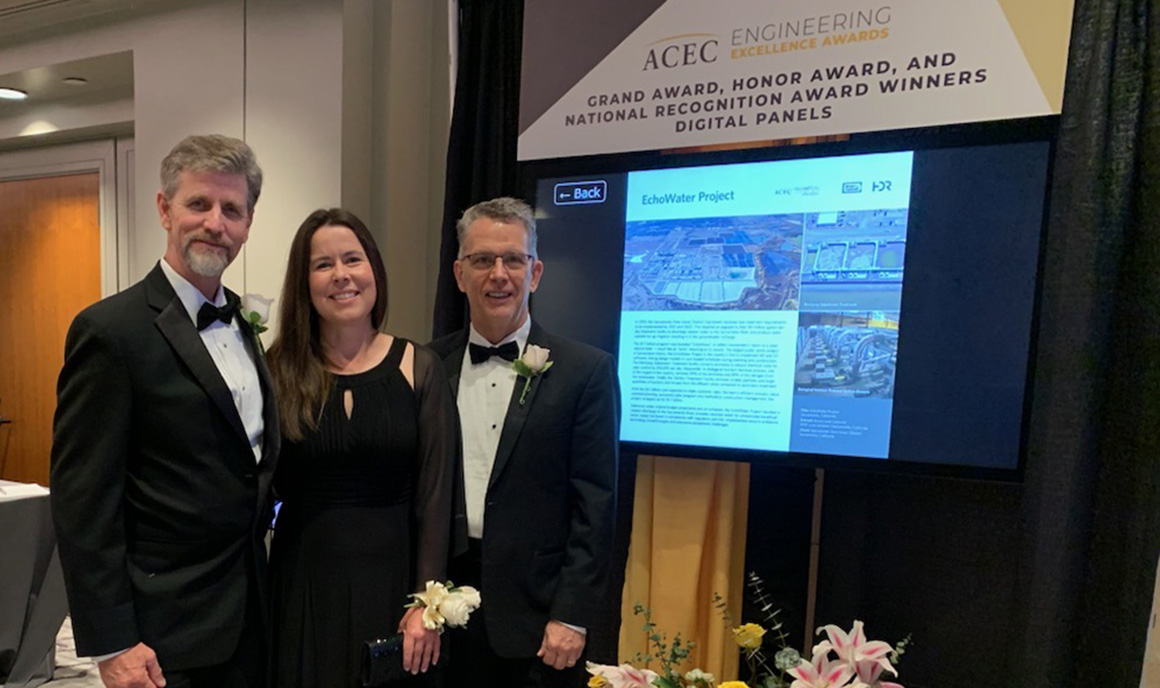 Brown and Caldwell staff of two men wearing black tuxedos and one woman in a long dark dress stand in front of a large screen showcasing Regional San's EchoWater Program, which won an ACEC 2024 Engineering Excellence Award.